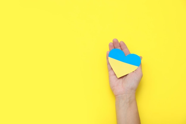 Woman holding paper heart in colors of Ukrainian flag on yellow background top view Space for text