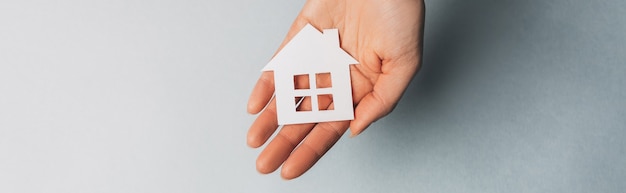 Woman holding a paper house with her hands with the sun on a light blue space