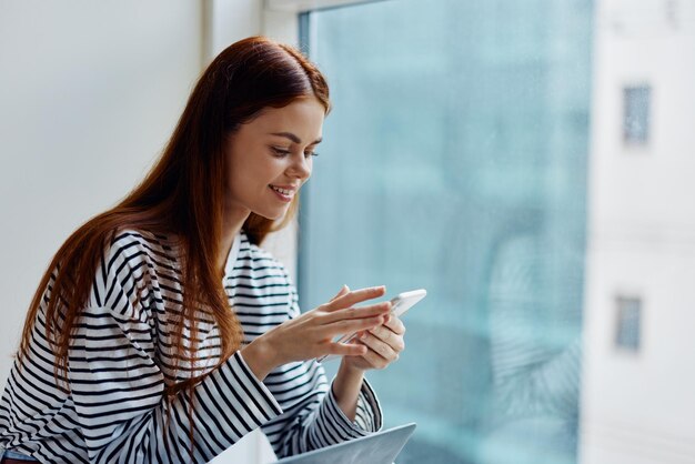 Photo woman holding a phone smile with teeth in front of a window overlooking the city the concept of working and learning online via the internet