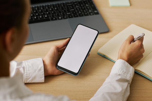 Woman holding phone with white screen mockup and taking notes at desk