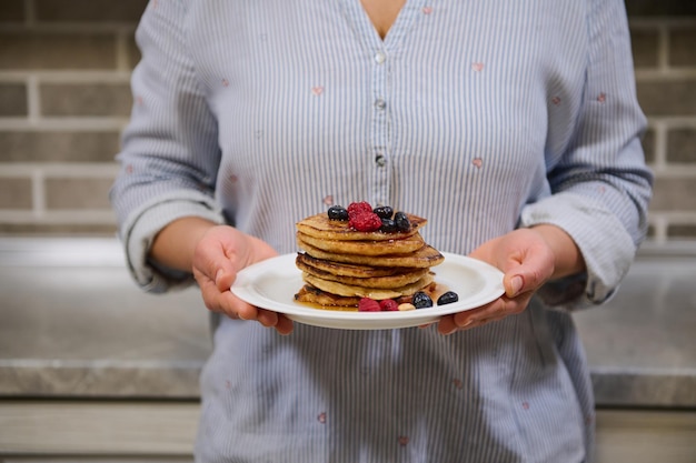 Woman holding plate of delicious homemade pancakes garnished with raspberries, blueberries and cashews, standing against kitchen countertop. Food for Shrovetide, Shrove Tuesday concept. Cropped image.