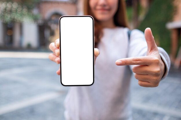 Woman holding and pointing finger at a mobile phone with blank white screen in the outdoors