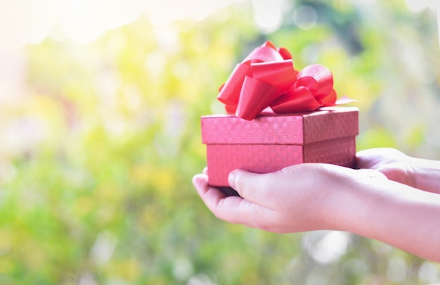 手に赤いプレゼントボックスを保持している女性を与える愛バレンタインデーコンセプト-自然の背景にリボンとギフトボックスを与える