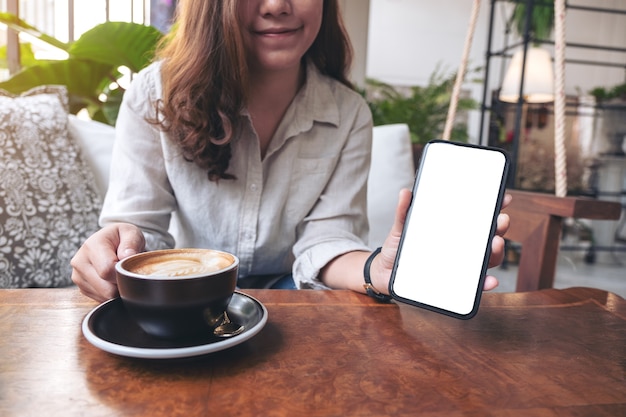 Photo woman holding and showing black mobile phone with blank white screen while drinking coffee in cafe