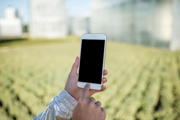 現代の水耕栽培温室の背景に空白の画面でスマートフォンを保持している女性