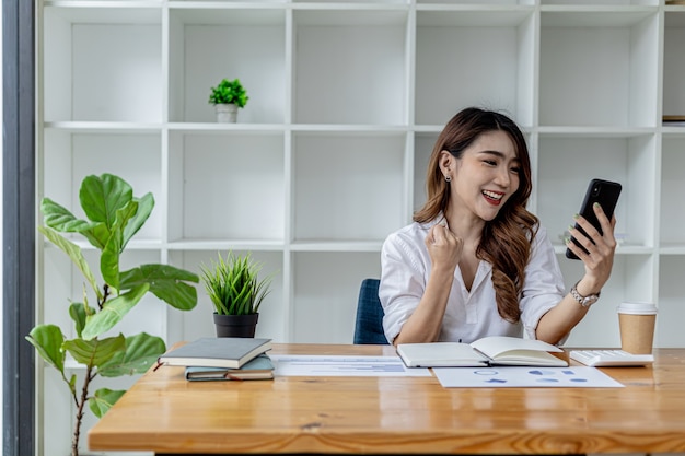 Woman holding a smartphone and gesturing, Asian business woman showing joy after watching the company's growing earnings in the past month, she manages the growth. Business administration concept.