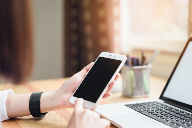 woman holding smartphone and using laptop on table in office room.