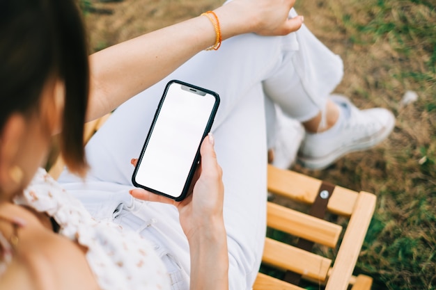 Woman holding a smartphone with a white screen mock up, sitting on a wooden chair outdoor in nature.