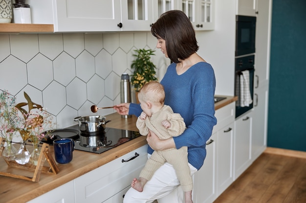 Woman holding spoon and watching something in saucepan. Young european mother holding newborn baby. Interior of kitchen in flat apartment. Concept of motherhood and child care