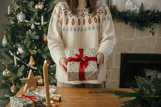 Woman holding stylish christmas gift with red ribbon at wooden table with festive decorations on background of decorated tree and fireplace in scandinavian room Merry Christmas