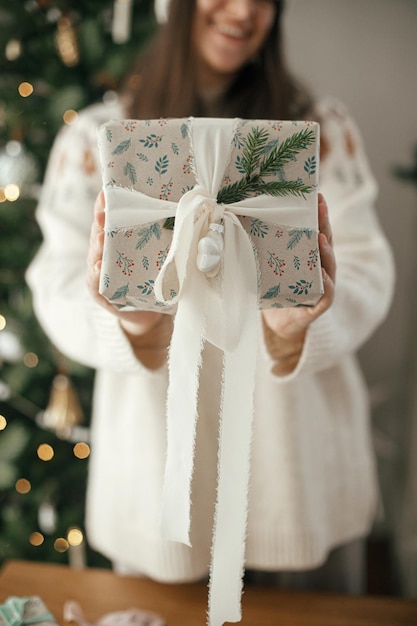 Женщина, держащая стильный рождественский подарок с лентой и еловой ветвью вблизи на фоне современного украшенного дерева в скандинавской комнате Счастливого Рождества и счастливых праздников
