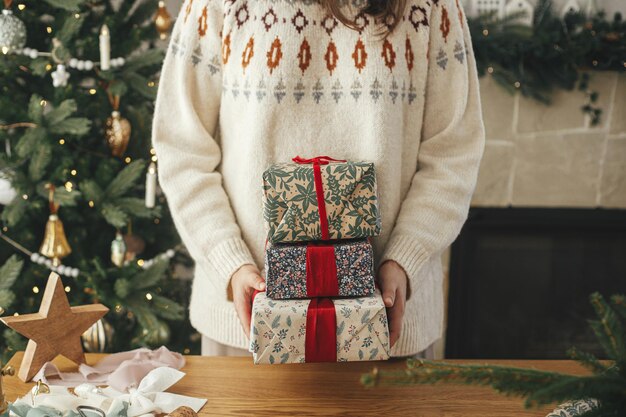 Photo woman holding stylish christmas gifts with red ribbon at wooden table with festive decorations on background of decorated tree and fireplace in scandinavian room merry christmas