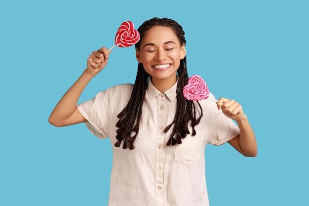 Woman holding two candies with heart shape keeps eyes closed has happy positive expression