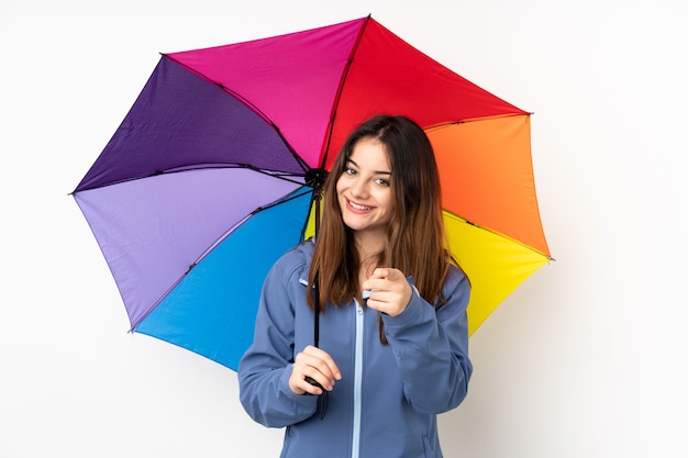 Woman holding an umbrella isolated on white wall points finger at you