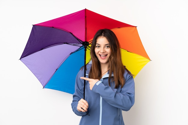 Woman holding an umbrella isolated on white wall surprised and pointing finger to the side