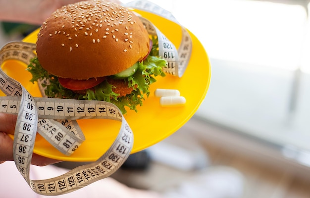 Woman holding unhealthy burger with measuring tape in kitchen