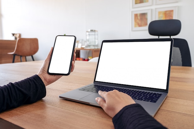 Una donna che tiene e utilizza il telefono cellulare mockup con schermo bianco vuoto e laptop sulla tavola di legno in ufficio