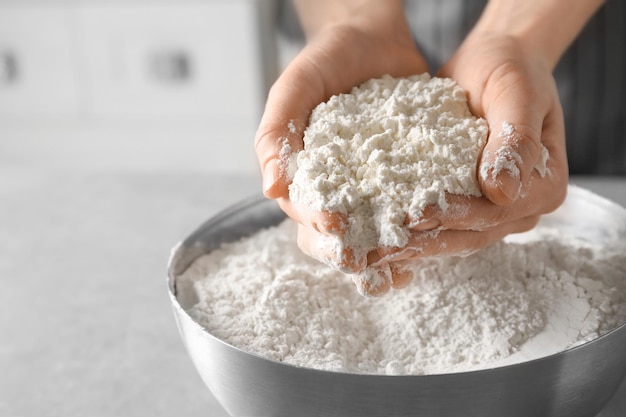 Woman holding wheat flour above bowl on table closeup