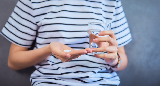 Woman holding white pill on hand and drinking water in glass on sofa in house