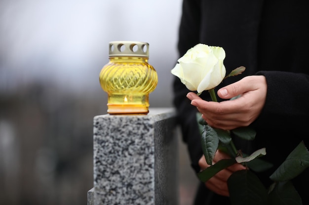 Woman holding white rose near grey granite tombstone with candle outdoors closeup Funeral ceremony