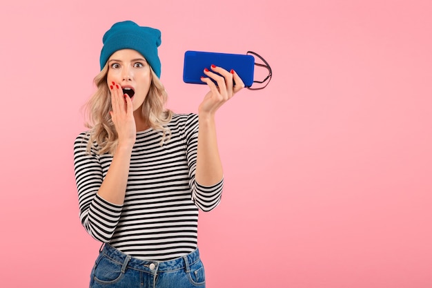 woman holding wireless speaker listening to music wearing striped shirt and blue hat smiling happy positive mood posing on pink