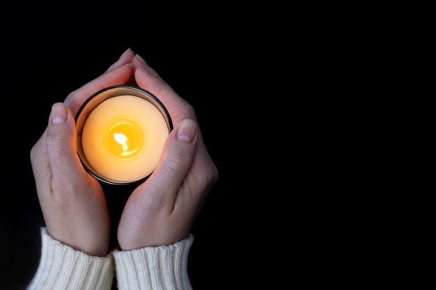 Photo woman holding with two hands a burning candle burning yellow flame close up on a black background