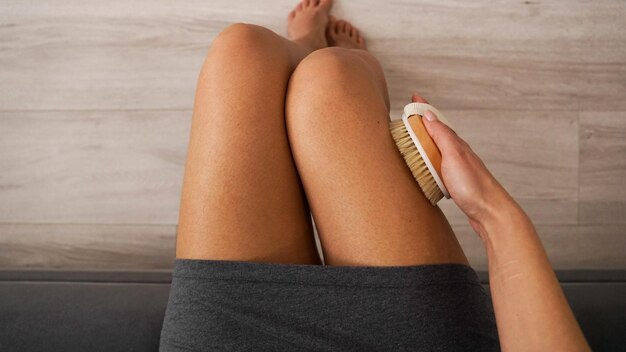 Woman holding wooden brush and doing dry massage on her legs self care concept