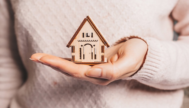 woman holding a wooden house with her hands, insurance on a light pink surface. sweet home 