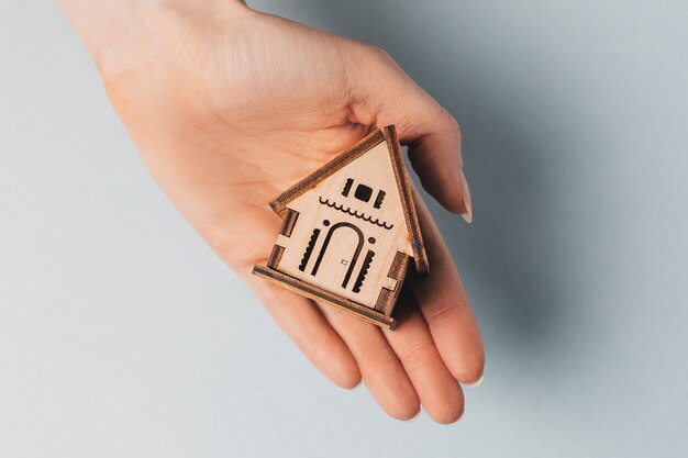 Woman holding a wooden house with her hands with the sun on a light blue background. Sweet home