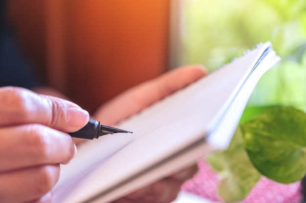 Женщина держит и пишет на блокноте перьевой ручкой, сидя в кафе с размытым зеленым фоном природы