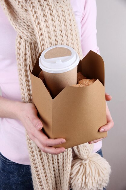 Woman holds box with coffee and cookies on grey background