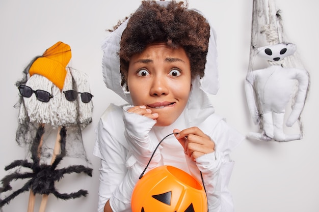 Photo woman holds carved pumpkin afraids of spooky creatures around bites lips dresses up in ghost cotume isolated on white