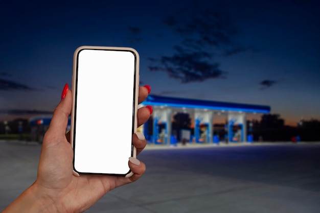 Woman holds a closeup of a smartphone with a white screen in his hands against the backdrop on a gas station Technology mockup for apps and websites