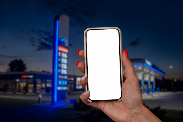 Woman holds a closeup of a smartphone with a white screen in his hands against the backdrop on a gas station Technology mockup for apps and websites