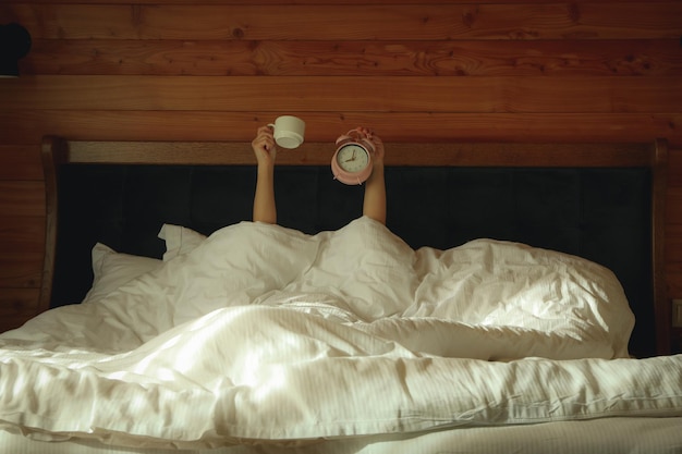 Woman holds cup and alarm and lying in bed