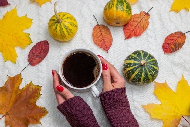 A woman holds a cup of coffee in front of autumn leaves and a pumpkin.