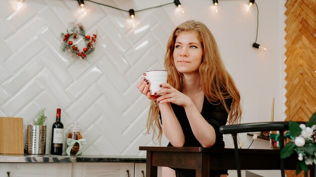 여자는 크리스마스 부엌에서 아침에 커피 한 잔을 들고 있습니다. 재미 있고 웃는 행복 한 젊은 여자