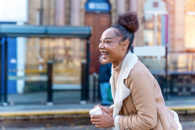 女性は一杯のコーヒーを保持しています トラムを待っている茶色のセーターで笑顔の巻き毛のブルネットの女性