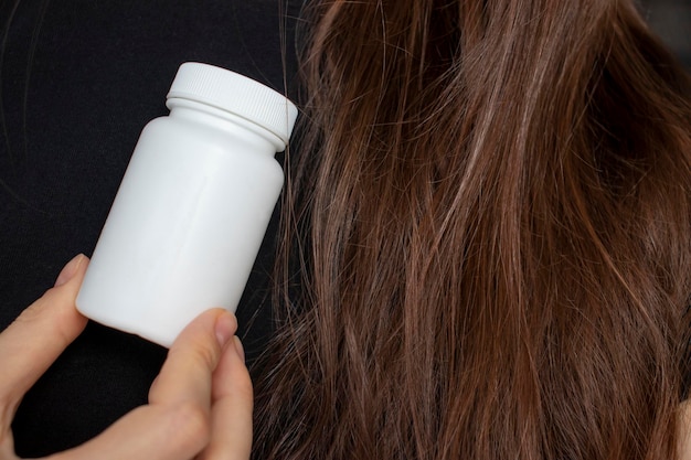 A woman holds an empty bottle of supplements and vitamins in her hand against a background of healthy hair and nails The concept of healthy hair care