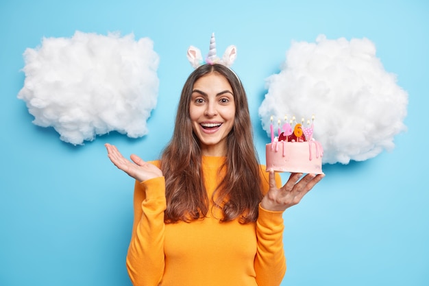 woman holds festive tasty birthday cake enjoys celebration raises palm poses on blue 