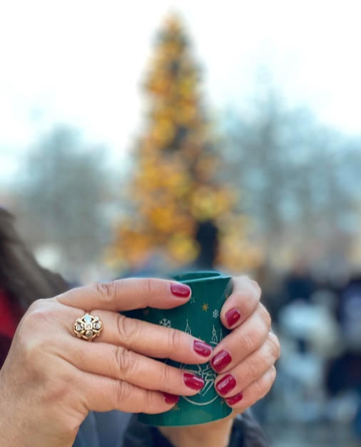 A woman holds a green cup in front of a christmas tree.