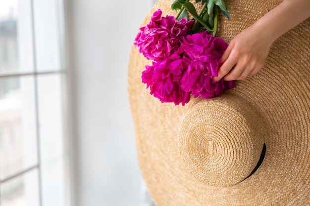 Woman holds in her hand a bouquet of peonies and a straw hat on a white background