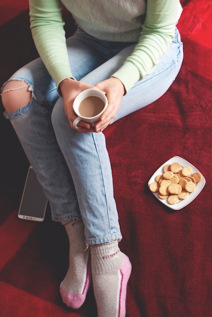 A woman holds in her hand a cup of coffee, small cookies next to her on a white saucer.