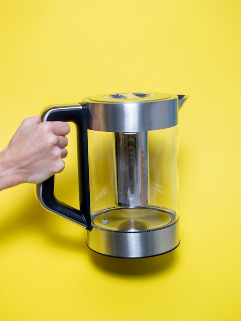 A woman holds in her hand a silver-colored electric metal kettle with various water heating modes on a bright yellow background.