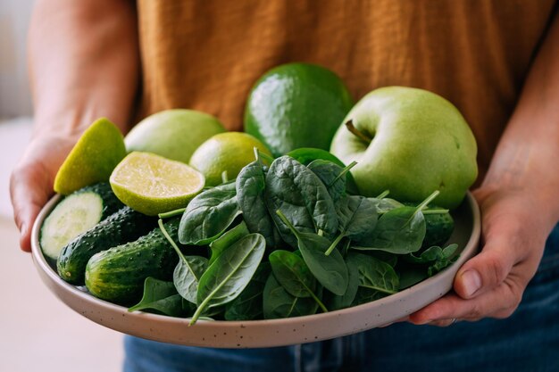 A woman holds in her hands fresh green vegetables and fruits in a large plate close-up soft focus