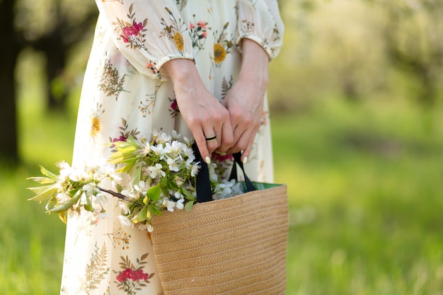 A woman holds in her hands a stylish wicker bag with blooming flowers in green park. Spring romantic mood and beuaty of nature