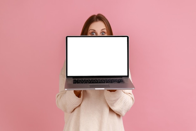 여자는 복사 공간이 있는 빈 디스플레이가 있는 노트북으로 얼굴의 절반을 덮고 있는 손에 노트북을 들고 있습니다.