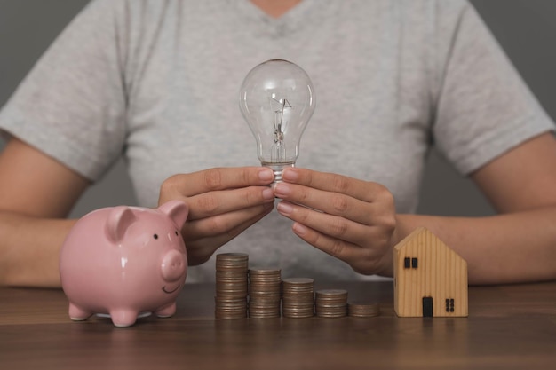 A woman holds a lightbulb and a stack of coins next to a piggy bank.