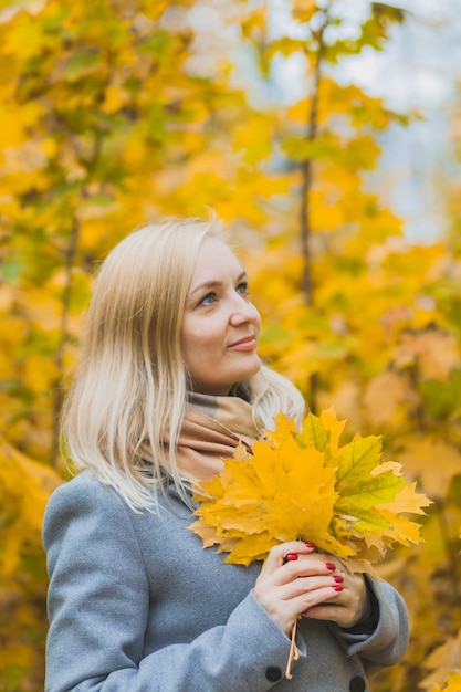 Женщина держит в руках кленовые желтые листья в парке на фоне деревьев