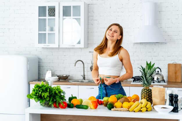 Woman holds measuring tape around waist Lot of fruits and vegetables on table Dieting